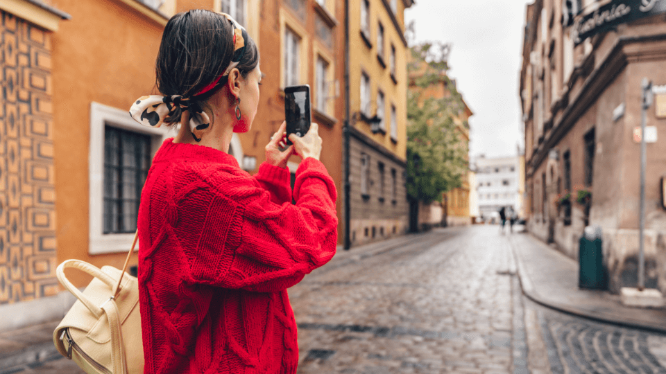 Mulher com um suéter vermelho e mochila bege fotografando uma rua de paralelepípedos cercada por prédios coloridos em um destino europeu.